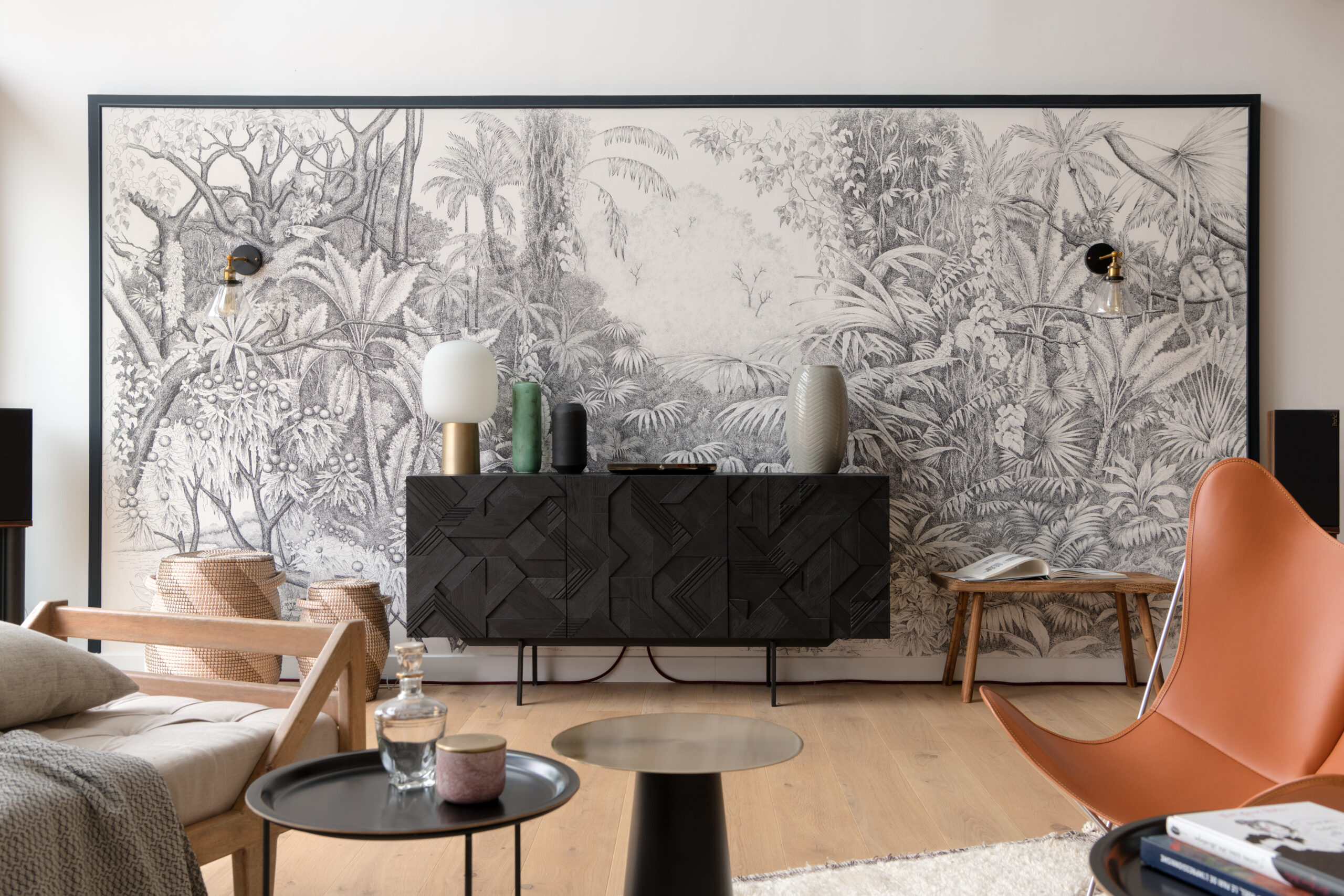 Salon avec un encadré de papier peint tropical noir et blanc. La pièce est composée de meubles en bois et de la couleur orange qui crée de beaux contrastes.